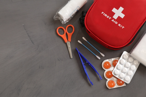 Flatlay mit Erste-Hilfe-Set. Zu sehen sind: ein weißer Verband, orange Schere, zwei Q-Tipps, blaue Pinzette, zwei Plister mit Tabletten.
