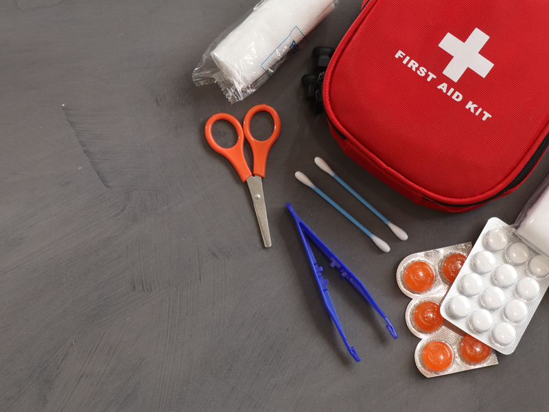 Flatlay mit Erste-Hilfe-Set. Zu sehen sind: ein weißer Verband, orange Schere, zwei Q-Tipps, blaue Pinzette, zwei Plister mit Tabletten.
