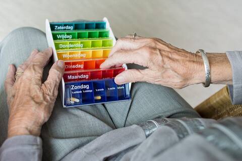 Eine ältere Frau, in grau gekleidet, hält ihre Tablettenbox, sortiert nach Wochentagen, in ihren Händen. 