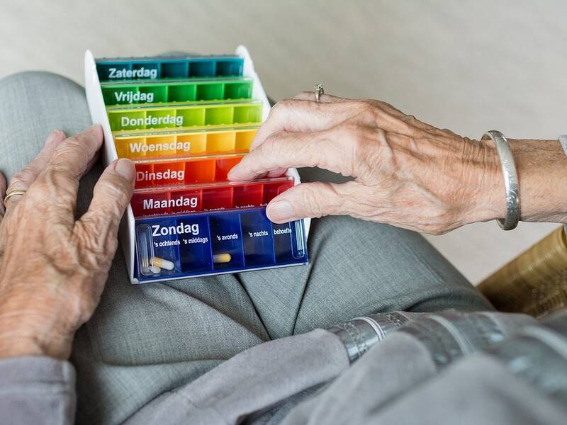 Eine ältere Frau, in grau gekleidet, hält ihre Tablettenbox, sortiert nach Wochentagen, in ihren Händen. 