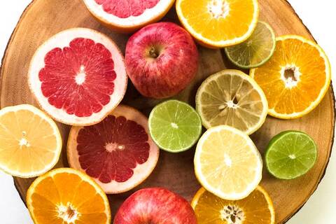Als Flatlay sind auf einer Holzscheibe verschiedene aufgeschnittene Früchte zu sehen: Zitrone, Orange, Limette, Apfel, Blutorange. Dazwischen liegen zwei rote Äpfel.