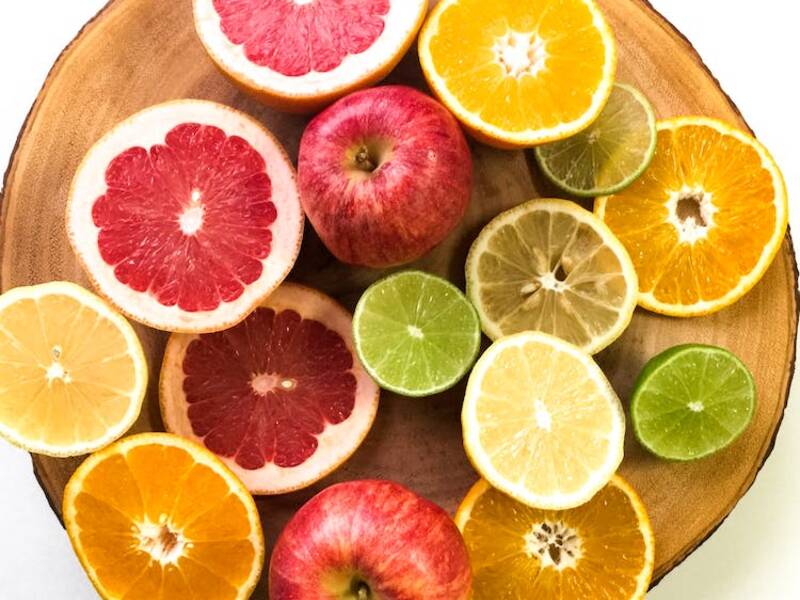 Als Flatlay sind auf einer Holzscheibe verschiedene aufgeschnittene Früchte zu sehen: Zitrone, Orange, Limette, Apfel, Blutorange. Dazwischen liegen zwei rote Äpfel.
