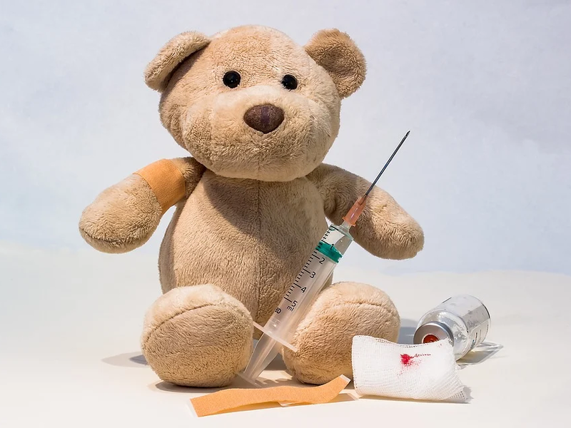 Ein hellbrauner Teddybär, mit einem Pflaster am rechten Arm. Vor ihm liegen ein Pflaster und ein Verband. In der Hand hat er eine Spritze.