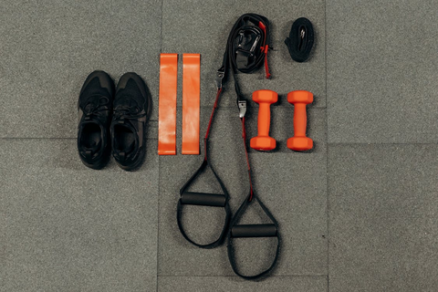 Flatlay zum Thema Sport. Untergrund sind graue Sportmatten. Darauf liegen von links nach rechts: ein Paar schwarze Sportschuhe, zwei orange Trainingsbänder, zwei schwarze Schlingen, zwei orange Hanteln