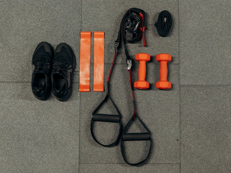 Flatlay zum Thema Sport. Untergrund sind graue Sportmatten. Darauf liegen von links nach rechts: ein Paar schwarze Sportschuhe, zwei orange Trainingsbänder, zwei schwarze Schlingen, zwei orange Hanteln