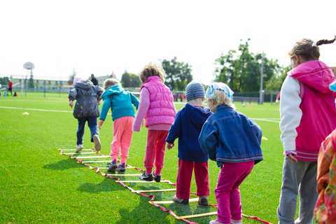 Sieben Kinder, in bunten Outfits, üben auf einem Rasensportplatz Springen mit einer Koordinationsleiter.
