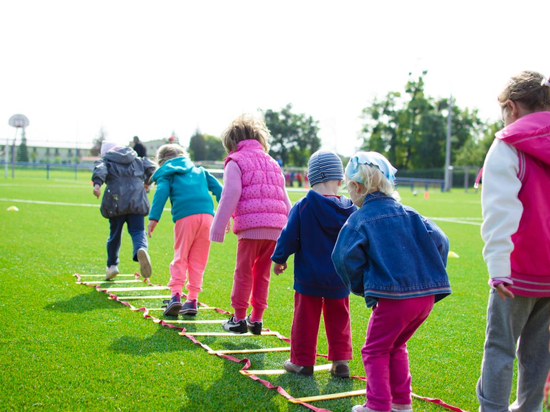 Sieben Kinder, in bunten Outfits, üben auf einem Rasensportplatz Springen mit einer Koordinationsleiter.