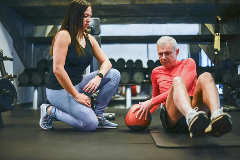 In einer Sporthalle sitzt ein älterer Herr ,in Begleitung einer persönlichen Trainerin, auf einer Sportmatte und macht eine Kräftigungsübung mit einem Ball.