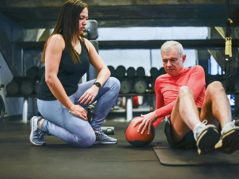 In einer Sporthalle sitzt ein älterer Herr ,in Begleitung einer persönlichen Trainerin, auf einer Sportmatte und macht eine Kräftigungsübung mit einem Ball.