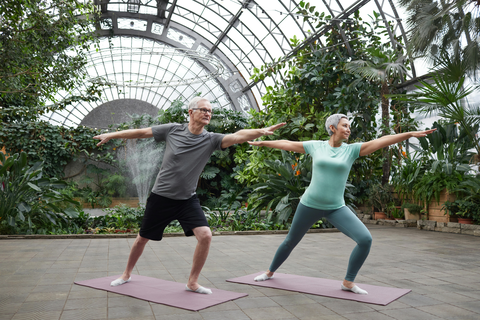 Ein Mann und eine Frau machen in einem großen Gewächshaus Yoga. Sie stehen auf altrosa-farbenen Yoga-Matten und haben die Yoga-Position Krieger inne.. Aussehen der Frau: kurze graue Haare, Leggings taupe, T-Shirt türkis. Aussehen des Mannes: kurze, graue Haare, Brille, kurze schwarze Hose, graues T-Shirt