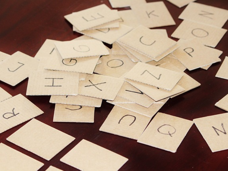 Auf einem braunen Tisch liegen weiße Papierschnipsel, auf denen in schwarzer Schrift einzelne Buchstaben des Alphabets stehen.