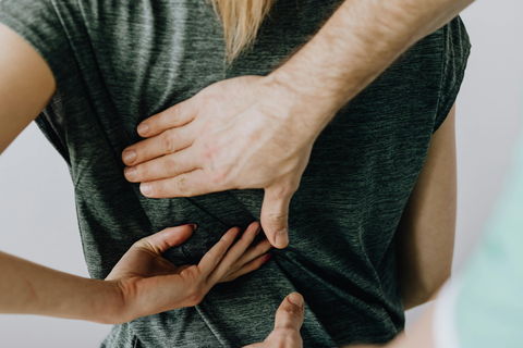 Rückansicht einer jungen auf Behandlungsliege sitzenden Frau (blondes Haar, zu Pferdeschwanz gebunden, grünes T-Shirt). Behandlung des Rücken durch Therapeuten (dunkles Haar, dunkle Brille)