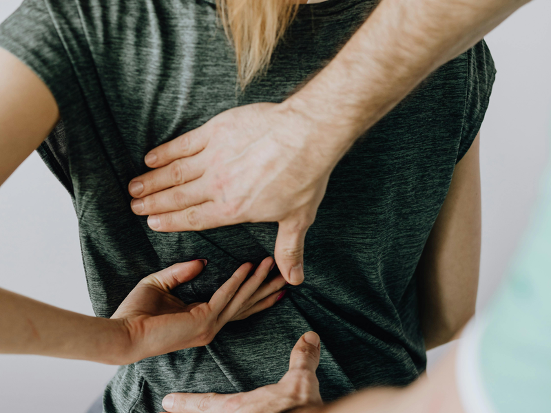 Rückansicht einer jungen auf Behandlungsliege sitzenden Frau (blondes Haar, zu Pferdeschwanz gebunden, grünes T-Shirt). Behandlung des Rücken durch Therapeuten (dunkles Haar, dunkle Brille)