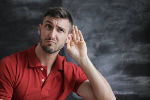 Es ist ein jüngerer Mann zu sehen (dunkle,kurze Haare, rotes T-Shirt). Er sitzt vor einem dunklen Hintergrund und hält sein Hand ans Ohr, als würde er etwas genauer hören wollen.