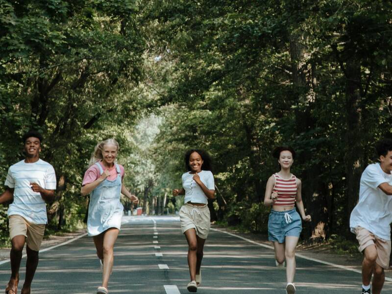 Sechs Jugendliche laufen, in unterschiedlichen sommerlichen Outfits, auf einer Allee der Kamera entgegen. 