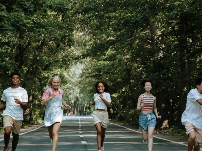 Sechs Jugendliche laufen, in unterschiedlichen sommerlichen Outfits, auf einer Allee der Kamera entgegen. 