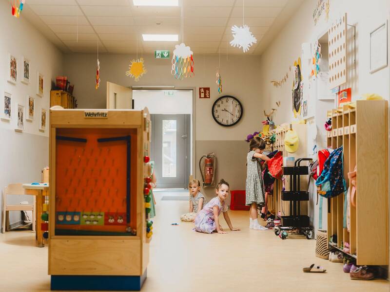 Blick in den Flur, ein Mädchen steht an der Garderobe und zwei weitere sitzen auf dem Boden daneben, eine Spielinsel steht in der Mitte des Flures