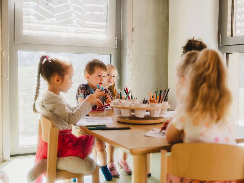 Kinder sitzen an einem Holztisch mit Malstiften und malen Bilder