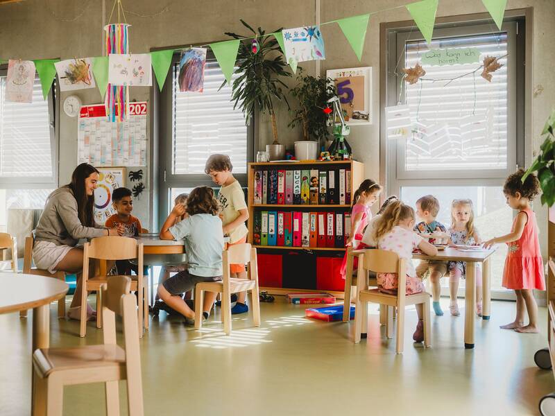 Blick in den Gruppenraum, an einem Holztisch sitzen 5 spielende Kinder und an einem zweiten Holztisch sitzt eine Erzieherin mit 4 Kindern und spielen zusammen