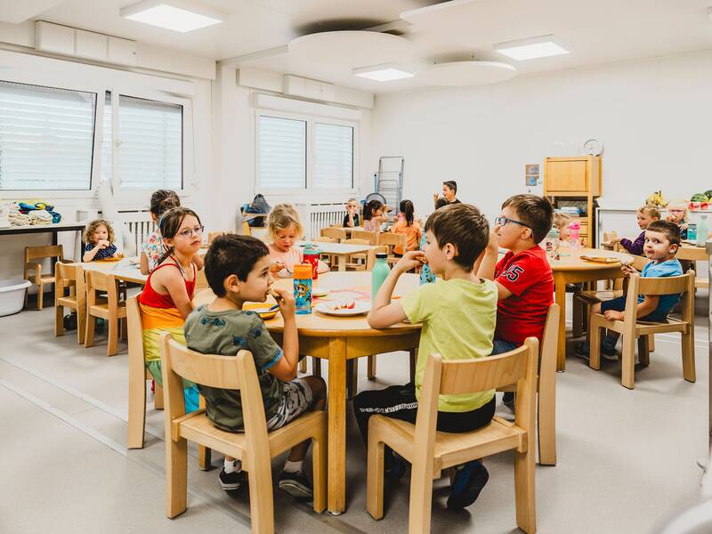 Blick in den Speisesaal in dem die Kinder an den Tischen sitzen und essen