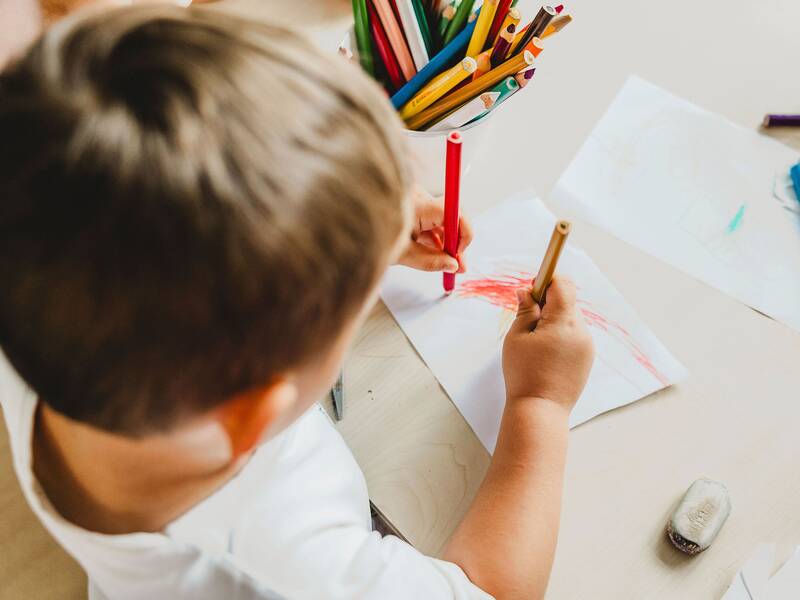 Blick von oben auf einen braunhaarigen Jungen der mit einem roten Stift in der linken und einem gelben Stift in der rechten Hand ein Bild malt