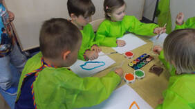 Vier Kinder in grünen Malerkitteln sitzen am Tisch und malen mit Wasserfarben.