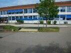 Außenansicht der Grundschule im Bisttal. Im Vordergrund ist ein Sandkasten, der mit einem blauen Sonnensegel überspannt ist.