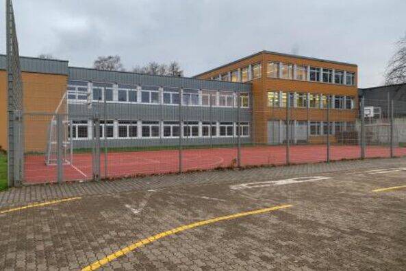 Aus Sicht des Parkplatzes ist der Standort Hostenbach der Bisttalschule Wadgassen Bous zu sehen. Vor dem Gebäude der Bisttalschule ist ein Multifunktionsfeld, mit zwei Basketballkörben und zwei Fußballtoren. Der Himmel ist bewölkt und regnerisch.