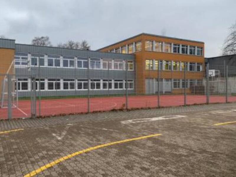 Aus Sicht des Parkplatzes ist der Standort Hostenbach der Bisttalschule Wadgassen Bous zu sehen. Vor dem Gebäude der Bisttalschule ist ein Multifunktionsfeld, mit zwei Basketballkörben und zwei Fußballtoren. Der Himmel ist bewölkt und regnerisch.