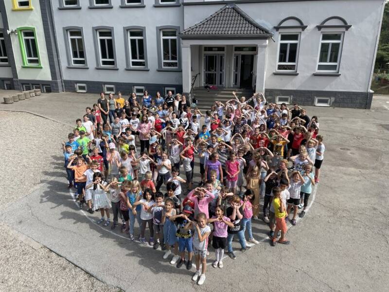 Die Schülerschaft hat sich auf dem Schulhof versammelt. Die Schüler:innen haben sich so aufgestellt, dass es aus der Vogelperspektive, wie ein Herz, aussieht.