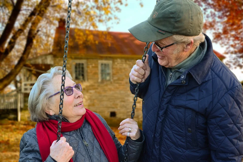 Eine ältere Frau (halblanges, graues Haar, Brille, roter Schal) sitzt auf einer Schaukel. Ein älterer Herr (Kappe, dunkle Brille) hält sich an der Schaukel fest und beugt sich zur Frau. Im Hintergrund sind Bäume zu sehen, die Herbstlaub tragen.