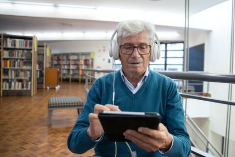 Ein älterer Herr (graues, kurzes Haar, dunkle Brille, hellblauer Pullover) sitzt in einer Bibliothek. Er hält ein Tablet in Händen und hat weiße Over-Ear-Kopfhörer an.