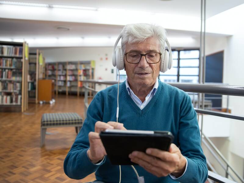 Ein älterer Herr (graues, kurzes Haar, dunkle Brille, hellblauer Pullover) sitzt in einer Bibliothek. Er hält ein Tablet in Händen und hat weiße Over-Ear-Kopfhörer an.