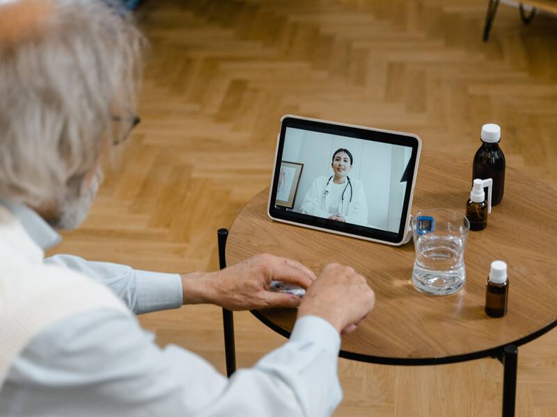 Ein älterer Herr nimmt in seinem Wohnzimmer eine telemedizinische Untersuchung wahr. Er sitzt auf einem Sofa, vor ihm  steht das Tablet auf dem Sofatisch. Auf dem Tablet ist die behandelnde weibliche medizinische Fachkraft zu sehen. 