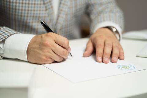 Eine Person unterzeichnet ein auf dem Tisch liegendes Dokument. Es sind nur die Hände und der Oberkörper der unterzeichnenden Person erkennbar.