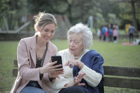Eine junge Frau und eine ältere Frau sitzen auf einer Parkbank. Die junge Frau zeigt etwas auf ihrem Smartphone, die ältere Frau hört interessiert zu. 