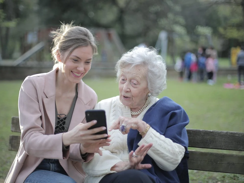 Eine junge Frau und eine ältere Frau sitzen auf einer Parkbank. Die junge Frau zeigt etwas auf ihrem Smartphone, die ältere Frau hört interessiert zu. 