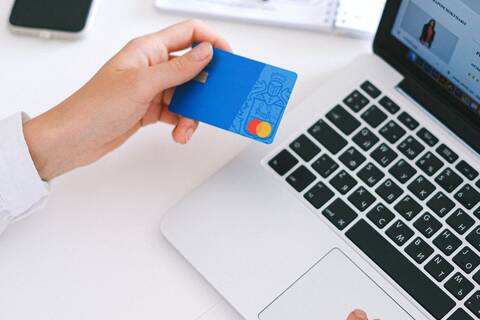 Eine Person shoppt online am Laptop. Sie hält eine Kreditkarte in den Händen. Links neben dem Laptop liegen ein Notizblock und ein Smartphone.