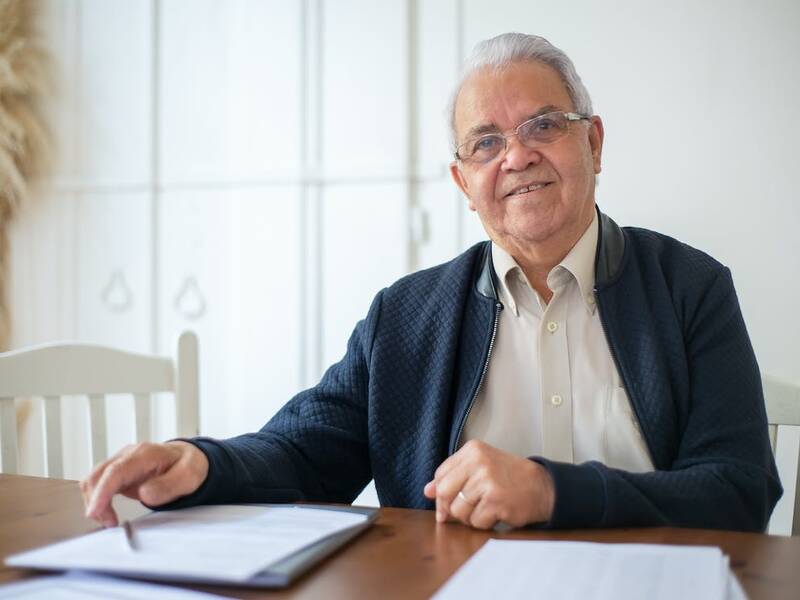 Ein älterer Herr (graues, welliges Haar, Brille) sitzt an einem braunen Tisch. Vor ihm liegen mehrere Dokumente.