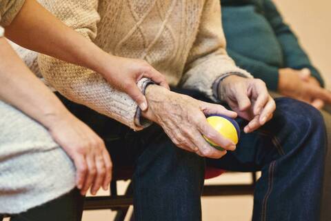 Eine ältere Person hält einen kleinen Ball in den Händen. Der Arm wird von einer jüngeren Person umfasst. 