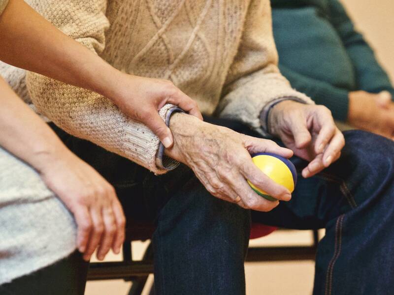 Eine ältere Person hält einen kleinen Ball in den Händen. Der Arm wird von einer jüngeren Person umfasst. 