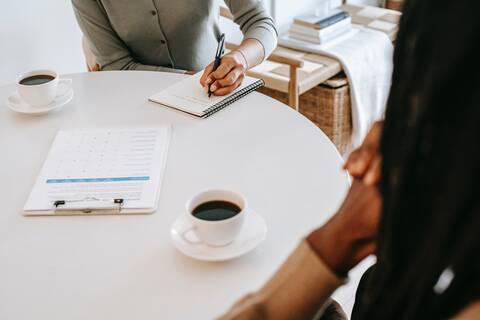 Es ist ein Tisch zu sehen, an dem zwei Personen sitzen, nur die Oberkörper und Arme sind sichtbar. Es stehen zwei gefüllte weiße Kaffeetassen auf dem Tisch. In der Mitte des Tisches liegt eine Checkliste. Eine der Personen schreibt auf einem Notizblock. 