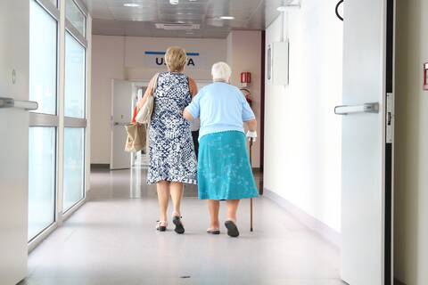 Krankenhausflur, in dem zwei Frauen in Rückansicht zu sehen sind. Die Frau, die links zu sehen ist, trägt an ihrem linken Arm zwei Taschen. An ihrem rechten Arm untergehakt ist eine ältere Frau, deren rechte Hand verbunden ist und die sich mit dieser auf einem Stock abstützt.